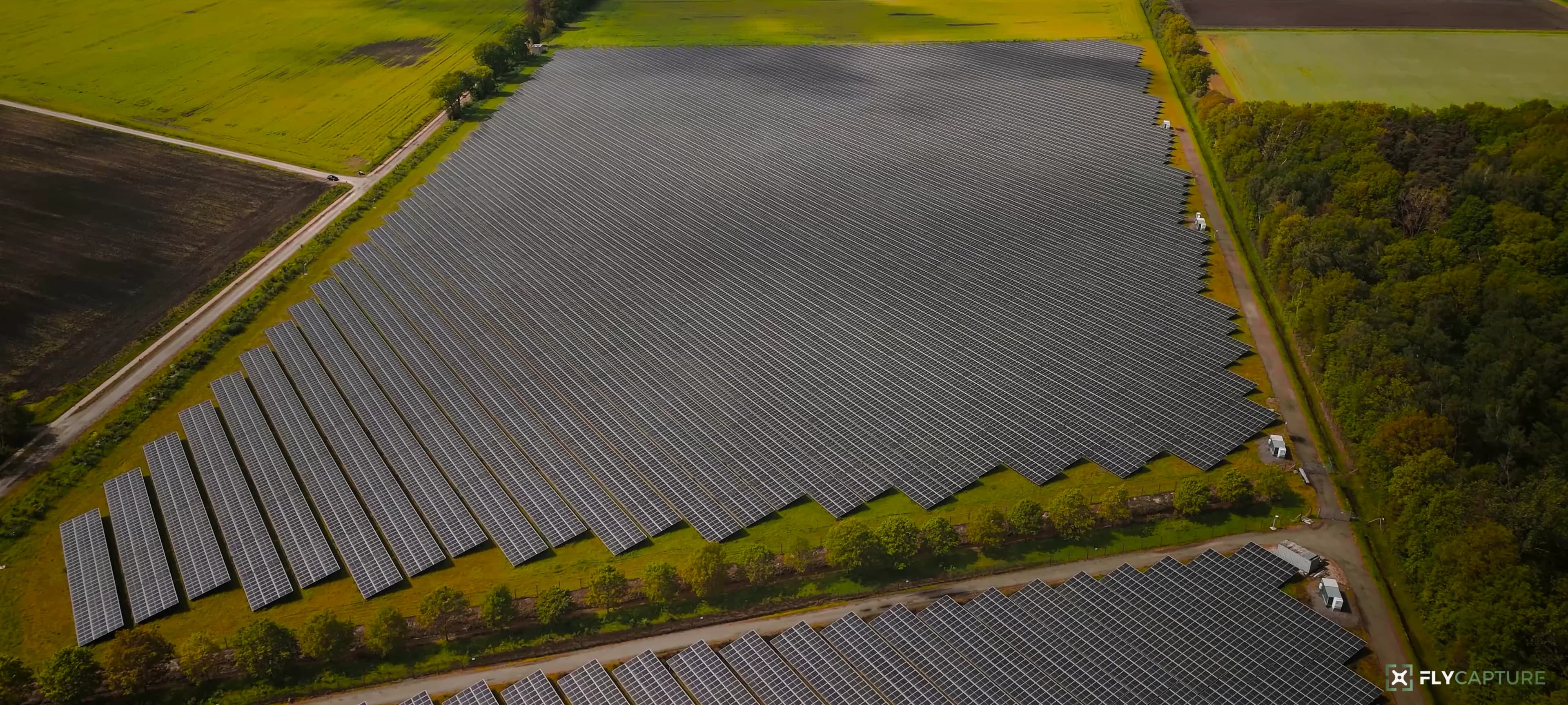 Zonnepark met lange rijen panelen tussen akkers en bos, vanuit de lucht